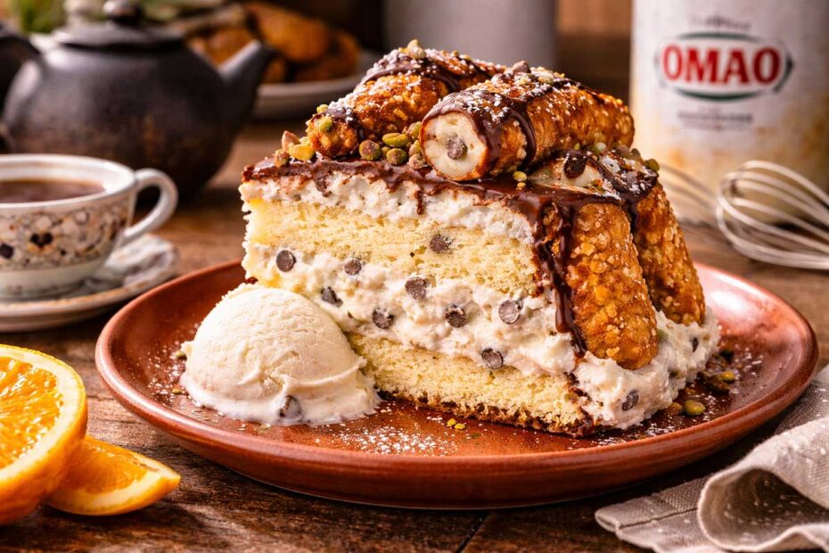Fatia generosa de Bolo de Cannoli com sorvete em um prato de louça rústica terracota. A fatia revela o recheio de ponto de fita com gotas de chocolate. A mesa de madeira maciça contém um bule de café fumegante, uma xícara de porcelana pintada à mão, cascas de laranja siciliana e um pacote de Açúcar União ao lado de um batedor de arame (fouet).