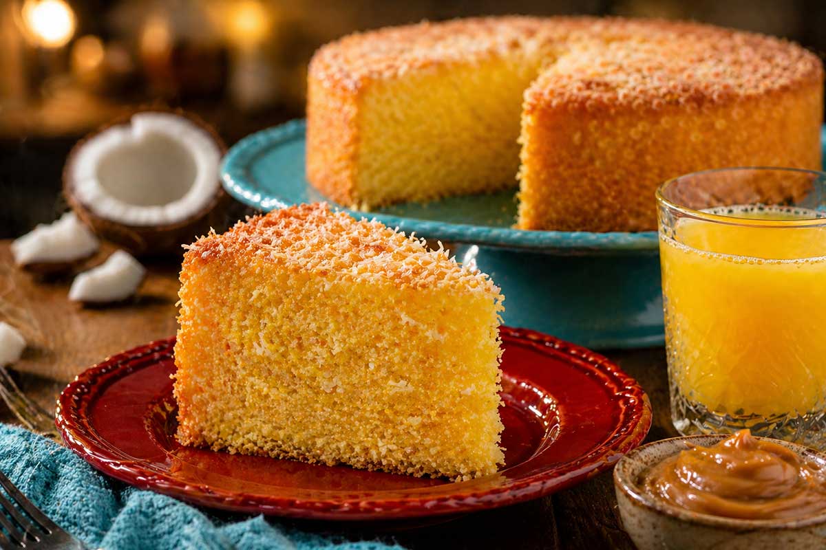 Close detalhado de uma fatia generosa de Bolo de Fubá com Coco em um prato de porcelana vermelha, destacando a massa aerada e amarela vibrante com fiapos de coco. A fatia está posicionada ao lado de um copo de suco de laranja natural e um pequeno pote com doce de leite cremoso. Ao fundo, o restante do bolo redondo de 20 cm permanece sobre a bailarina azul, criando um contraste cromático moderno e sofisticado em uma cozinha de alto padrão com iluminação quente.