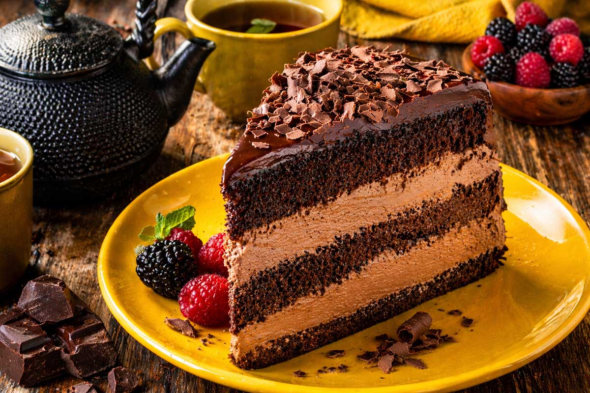 Uma fatia generosa de bolo de chocolate de frigideira servida em um prato de ceramica amarela vibrante sobre uma mesa rustica de madeira de fazenda. A fatia revela camadas intercaladas de massa acetinada umedecida com leite e um recheio de chantilly de chocolate firme e aerado. Ao redor estao dispostos um bule de cha de ferro, xicaras de cha de hortela, pedaços de chocolate rustico e frutas frescas como amoras e framboesas. A iluminacao lateral destaca a umidade residual da massa e o contraste cromatico entre o marrom do recheio e o amarelo do prato ativa as glandulas salivares.