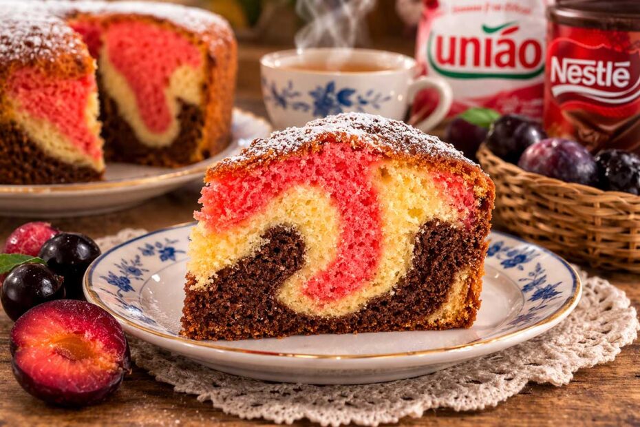 Fatia de bolo mesclado napolitano com massa aerada em tons de rosa, marrom e marfim, servida em prato de porcelana sobre toalha de renda. Ao fundo, xícara de café fumegante, ameixas frescas e embalagens de açúcar União e chocolate Nestlé em mesa de madeira rústica.