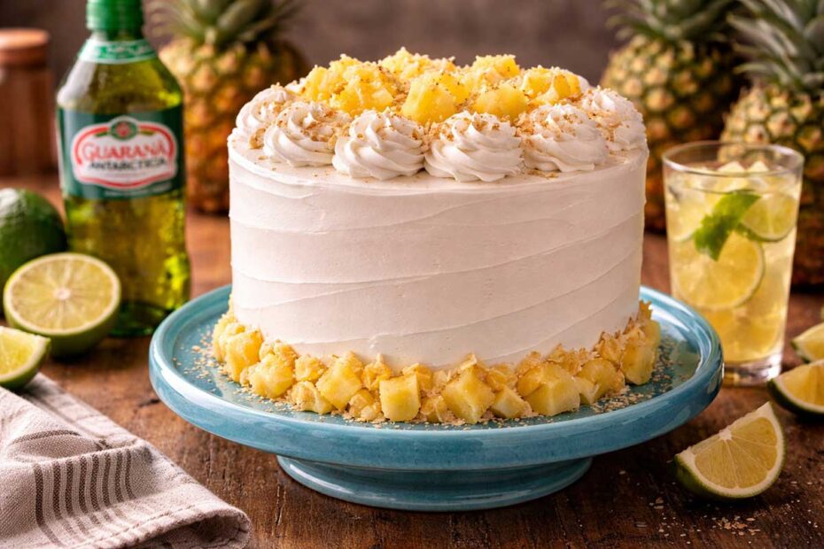 Bolo de Aniversário de Abacaxi alto e imponente, decorado com chantilly branco em bicos de flor e pedaços de abacaxi dourados no topo, sobre suporte de cerâmica azul vibrante em mesa rústica de madeira com bule de café fumegante, xícara de porcelana e um pacote de Açúcar União ao fundo.