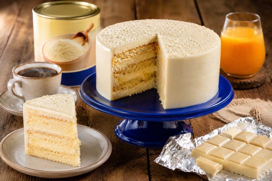 Fotografia gastronômica em ângulo frontal de um Bolo de Leite Ninho redondo, com 20 cm de diâmetro e altura de Tall Cake, posicionado sobre uma bailarina de confeitaria azul cobalto vibrante que contrasta com a cobertura branca acetinada. O bolo está fatiado, revelando camadas internas úmidas e recheio cremoso. A cena de mesa posta rústica sobre madeira inclui uma fatia em prato de cerâmica, uma xícara de café fumegante, um copo de suco de laranja natural, uma barra de chocolate branco aberta em papel alumínio e uma lata de leite em pó ao fundo com uma colher de madeira.